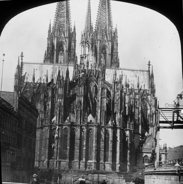 Ein Schwarz-Weiß-Foto der Kölner Dom in Köln, Deutschland, mit einer hohen Spitze, umgeben von anderen Gebäuden, Menschen, die davor gehen, und einer Brücke auf der rechten Seite.