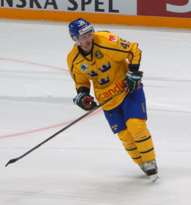Eishockeyspieler in Bewegung auf dem Eis mit Helm, Handschuhen und Schlittschuhen, der einen Hockeystock hält, mit einem textgefüllten Board im Hintergrund.