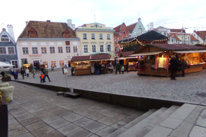 Ein lebendiger Weihnachtsmarkt in Tallinn, Estland, mit Menschen um geschmückte Stände, Gebäuden mit Fenstern im Hintergrund, bewölktem Himmel, Treppe, die zum Markt führt, und Töpfen mit Pflanzen drumherum.