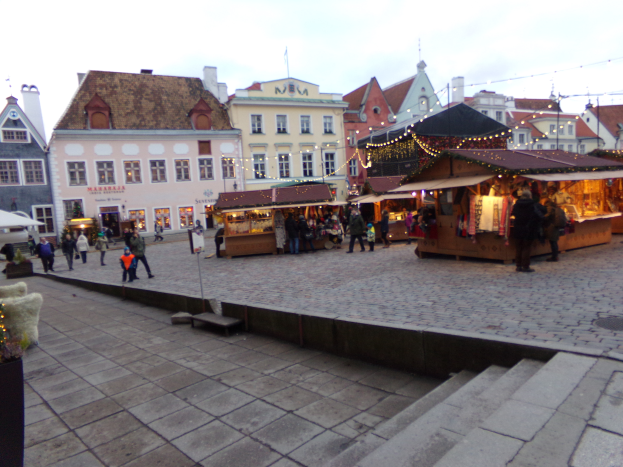 Ein lebendiger Weihnachtsmarkt in Tallinn, Estland, mit Menschen um geschmückte Stände, Gebäuden mit Fenstern im Hintergrund, bewölktem Himmel, Treppe, die zum Markt führt, und Töpfen mit Pflanzen drumherum.