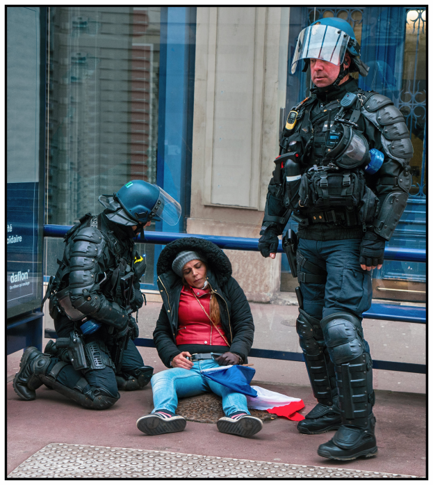 Eine Frau sitzt neben zwei uniformierten Polizeibeamten mit Helmen und Schutzkleidung auf dem Boden. Im Hintergrund ist ein Gebäude mit Glastüren und einer Texttafel zu sehen.