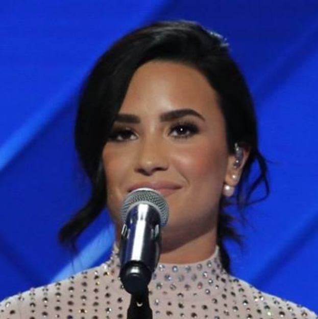 Demi Lovato steht selbstbewusst auf der Grammy-Bühne vor einem Mikrofon, tr├Ągt ein schwarzes Kleid und einen glatten Bob, mit einem blauen Hintergrund.