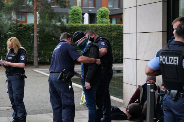 Polizisten vor einem Gebäude mit einer Frau, die ein Buch und einen Stift hält, während Menschen in der Nähe sitzen und stehen, mit Bäumen und Gebäuden im Hintergrund.