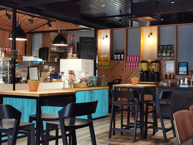 Innenansicht eines Restaurants mit Holztischen, Stühlen, einer Topfpflanze auf einem Tisch, einer Tafel, einem Kühlschrank, einem Feuerlöscher, Regalen mit Flaschen, Schränken mit Gegenständen, einem Foto auf der Wand, Deckenlampen und Lampen.