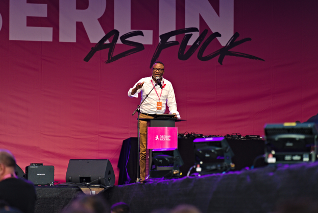 Ein Mann in einem weißen Hemd und braunen Hosen steht auf einer Bühne und hält ein Mikrofon, mit einem pinken Banner dahinter, das 'Berlin as Fuck' lautet, und Lautsprechern auf der Bühne.