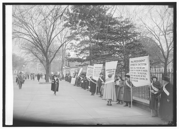 Eine Gruppe von Suffragetten marschiert auf einem baumbestandenen Bürgersteig, hält Schilder, mit einem Zaun auf der rechten Seite, dargestellt in Schwarz-Weiß.