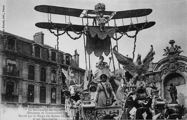 Ein Schwarz-Weiß-Foto eines Umzugswagens mit Statuen und einem Flugzeug, der eine Gruppe von Menschen trägt, mit einem Gebäude und Himmel im Hintergrund und einem Text am unteren Rand, der darauf hinweist, dass es ein Foto des Pariser Karnevals ist.