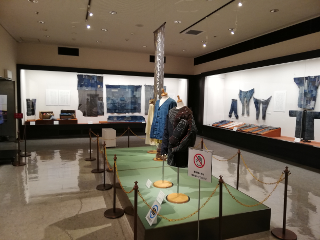 Ein Raum voller Schaufensterpuppen in traditioneller japanischer Kleidung, Vitrinen mit verschiedenen Kleidungsstücken, Informationsschilder, Pfosten mit Ketten und Deckenleuchten.