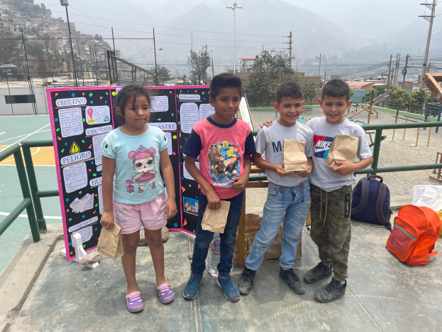 Eine Gruppe von Kindern steht auf einem Basketballfeld, jeder hält einen Papiertüte, mit Tüten und Gegenständen rechts, einer Texttafel links und einem Hintergrund aus Zaun, Pfosten, Straßenlaternen, Strommasten mit Drähten, Bäumen, Gebäuden, Hügeln und dem Himmel.