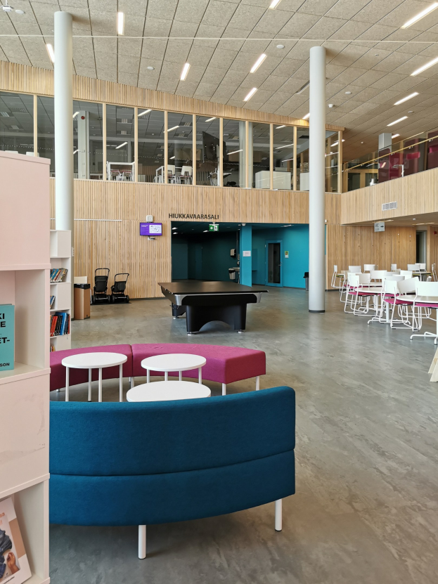 Ein geräumiger Raum, der einer Schulbibliothek ähnelt, mit mehreren Tischen, Stühlen, Sofas, Hockern, Bücherregalen, Deckenleuchten, einem wandmontierten Fernseher und strukturellen Säulen.