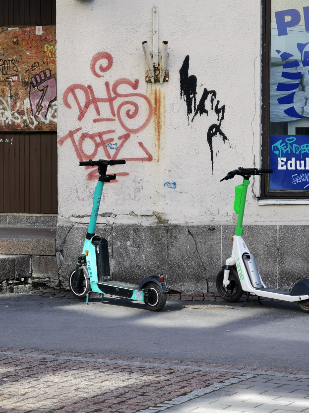 Zwei elektrische Roller nebeneinander auf einer Straße geparkt mit einer Graffiti-bedeckten Wand und einem Plakat im Fenster im Hintergrund.