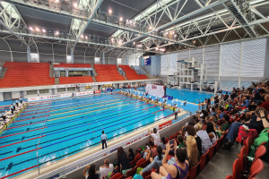 Großer Hallenbad mit vielen Menschen auf Stühlen drumherum und einigen im Wasser stehend, mit einer Decke mit Lampen und Eisenstangen und Schildern im Hintergrund.