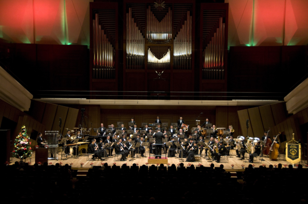 Großes Orchester auf der Bühne mit Weihnachtsbaum im Hintergrund, Musiker auf Stühlen sitzend, Dirigent am Podium und Texttafeln rechts.