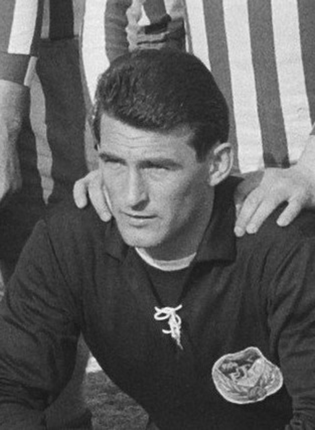 Ein Schwarz-Weiß-Foto eines Mannes, der auf einem grünen Fußballfeld sitzt und ein T-Shirt und eine Kette trägt, mit zwei Personen dahinter.