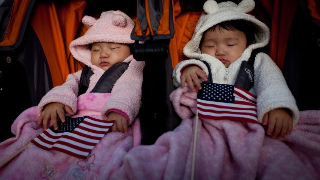Zwei Babys schlafen in einem orange-schwarzen Babywagen mit weißen, roten und blauen Flaggen und pinken Decken unten.