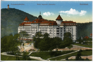 Eine alte Postkarte zeigt das Hotel Imperial in Karlsbad, Deutschland, ein großes Gebäude mit zahlreichen Fenstern, umgeben von Bäumen, Pflanzen und Gras, mit einem Weg im Vordergrund und Hügeln und einem bewölkten Himmel im Hintergrund, und Text oben auf dem Bild.