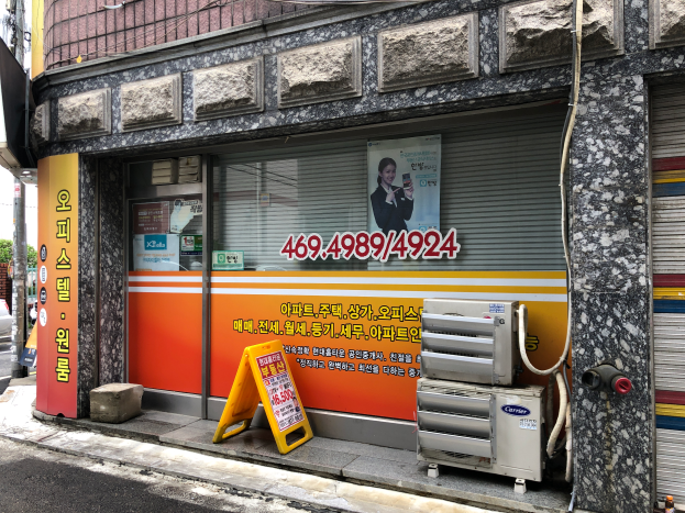 Ein Gebäude mit einer Tafel, die "Koreanische Klimaservice" anzeigt, einem Rollladen, einer Tafel mit Text und einem Personenbild, Geräten auf dem Boden, Pfosten, Bäumen, einem Auto auf der Straße und Drähten unter einem bewölkten Himmel.