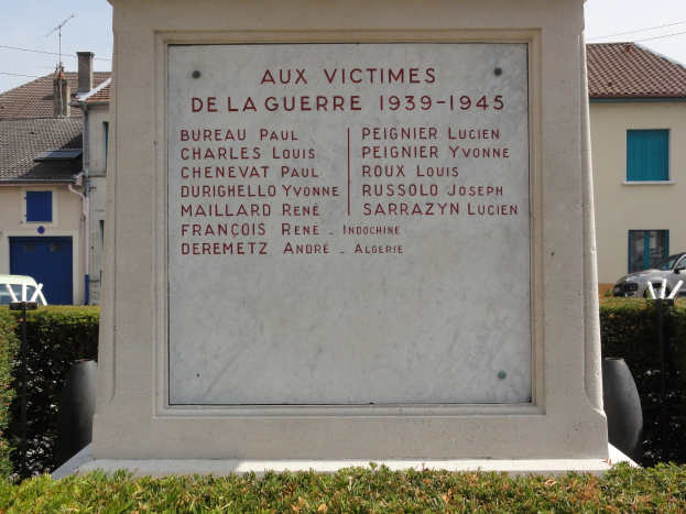 Ein Denkmal für die Opfer des Krieges von 1939–1945 steht vor einem Gebäude mit der Aufschrift "Aux Victimes de la Guerre 1939-1945", umgeben von Pflanzen, Fahrzeugen, Strommasten, Leitungen und nahegelegenen Häusern unter einem bewölkten Himmel.
