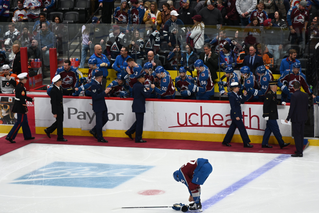 Ein Hockey-Spieler auf dem Eis mit einem Stock in der Hand, umgeben von einer Gruppe von Menschen mit Kappen und Hockey-Stöcken, mit Zuschauern und Gittern, auf denen "Colorado Avalanche vs San Jose Sharks - 3/24/15 NHL Pick, Odds, and Prediction" steht.