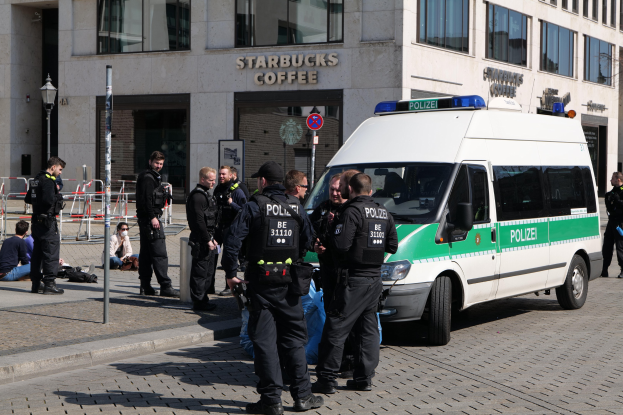 Eine Gruppe von Polizeibeamten steht vor einem Starbucks-Café, rechts ist ein Van und links ein paar Menschen zu sehen; im Hintergrund ist ein Gebäude mit Fenstern, ein Schild, ein Laternenmast und ein Zaun zu erkennen.