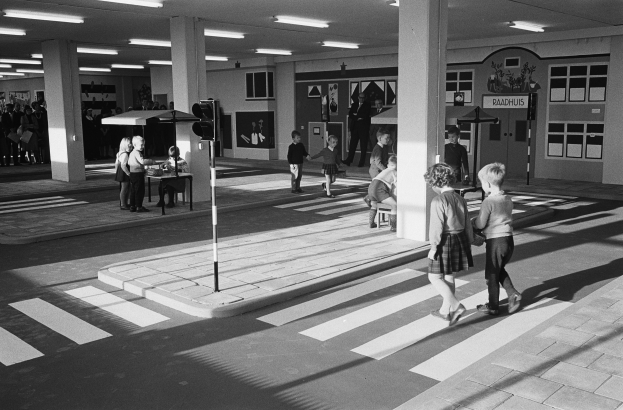 Schwarzes und weißes Foto von Kindern, die eine Straße an einer Ampel überqueren, mit Fußgängern und Bänken im Vordergrund und Säulen mit Deckenleuchten im Hintergrund.