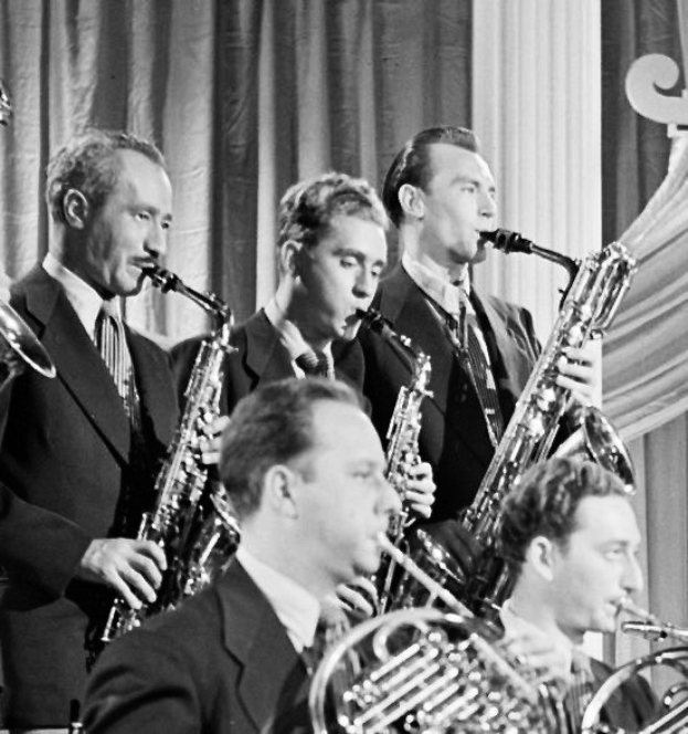 Eine Gruppe von Männern in Blazern und Krawatten, die Saxophone spielen, vor einem Vorhang abgebildet in Schwarz-Weiß.