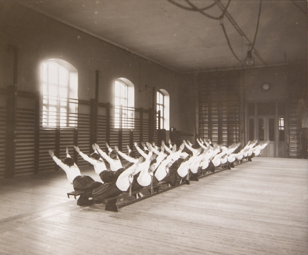 Gruppe von Menschen, die Pilates-Übungen in einem Fitnessstudio mit Fenstern und einer Uhr an der Wand im Hintergrund durchführen.