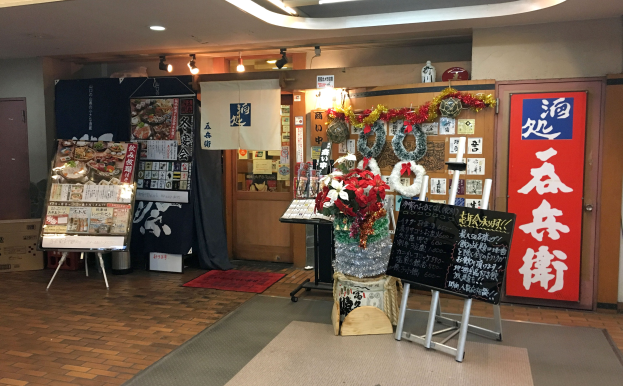 Eingangsbereich eines japanischen Restaurants mit Regalen, auf denen Gegenstände ausgebreitet sind, einer Vase mit Blumen, einer Tafel auf einem Ständer, einem Teppich, einer Pappkiste, Papieren an der Wand, Deckenlampen, einem Vorhang, einer Tür und einem Schild auf der rechten Seite.