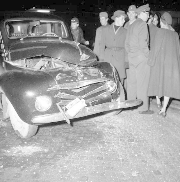 Schwarzes und weißes Bild eines schwer beschädigten Autos am Straßenrand, mit einer Gruppe von Menschen, die es inspizieren.