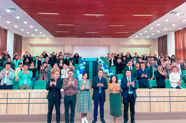 Eine Gruppe von Menschen steht vor einer Menge, einige klatschen, in einem Raum mit Stühlen, Tischen, Ballons, Vorhängen und Deckenleuchten, feiern die Eröffnung einer neuen Schule.