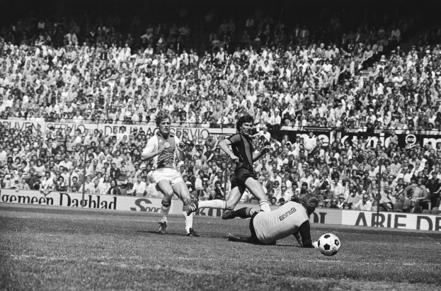 Ein Schwarz-Weiß-Bild einer Gruppe von Männern, die auf einem Feld Fußball spielen, mit Zuschauern im Hintergrund und einer Fahne mit der Aufschrift "Bundesliga 1978-79 Ajax 1-1 Hannover 96".