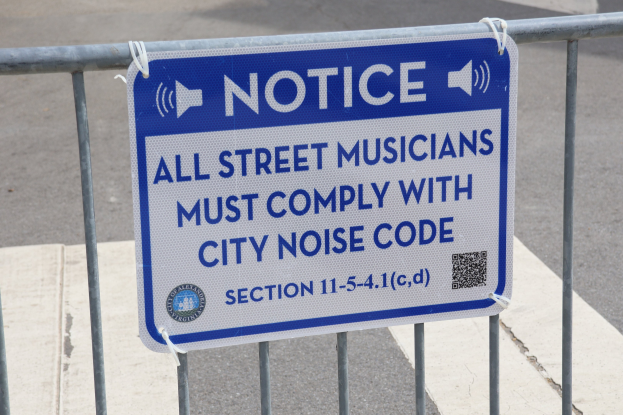 Anschlagtafel an einem Zaun mit einem Schild, das Straßenmusikern die Einhaltung der städtischen Lärmvorschriften auferlegt.