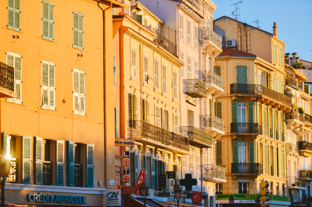 Eine Stadtstraße mit hohen Gebäuden, die Fenster, Balkone und Schildertafeln aufweist und von Bäumen und Straßenlaternen gesäumt ist, unter einem klaren blauen Himmel.
