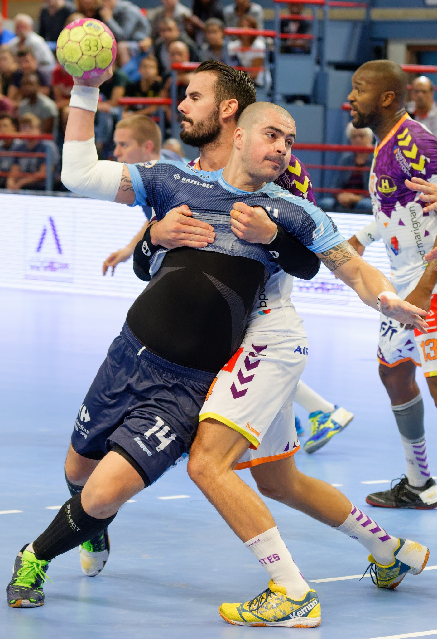 Männer beim Handballspielen in einer Stadion, einer hält den Ball, mit Zuschauern auf Stühlen, Geländern und einer Wand im Hintergrund.