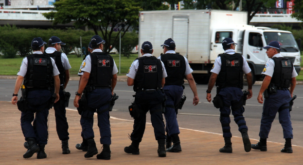 Eine Gruppe von Beamten in Uniform, einschließlich Mützen, Hemden, Schutzwesten, Uhren, Hosen und Schuhen, die auf einer Straße mit einem geparkten Lastwagen auf der rechten Seite neben einem Zaun gehen. Im Hintergrund sind Gebäude, Bäume, Pflanzen, Pfosten und Banner zu sehen, mit einer Brücke in der oberen rechten Ecke.