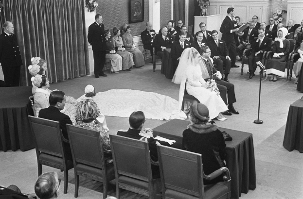 Schwarz-weiß-Foto einer Hochzeitszeremonie mit Sitzenden und Stehenden, einem Mann mit Mikrofon, einer Frau mit Blumen, einem Podium mit Mikrofon, Blumenschmuck, einem Vorhang, einem gerahmten Foto an der Wand und einer Tür im Hintergrund.