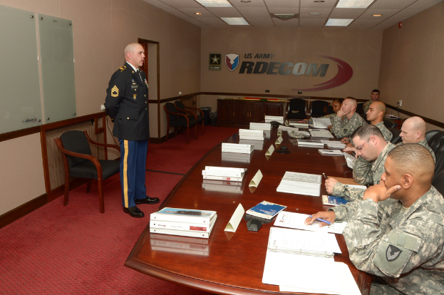 Ein Schreibtisch mit Papieren und Akten, auf dem rechten Sitzende in Uniform und links ein stehender Person, dahinter ein Stuhl und "US Army RDECOM" auf der Rückwand.