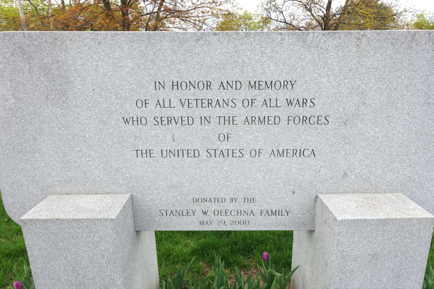 Gedenkstein mit einer Inschrift für Veteranen der US-Streitkräfte, umgeben von Pflanzen, Blumen, Gras, Bäumen und einem bewölkten Himmel.