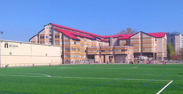 Fußballfeld mit einem großen Gebäude, das San Visitor Academy im Hintergrund zeigt, umgeben von Bäumen und Laternen, einige Menschen auf dem Feld und Himmel darüber.