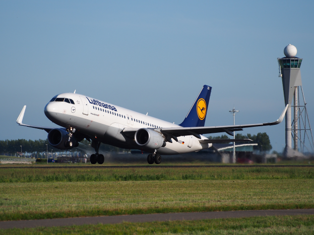Ein Lufthansa Airbus A320-200 Flug von Frankfurt Airport, mit grünem Gras und einer Straße im Vordergrund und hohen Türmen, Bäumen und einem klaren blauen Himmel im Hintergrund.