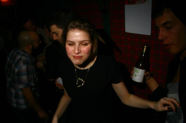Eine Gruppe von Menschen in einem Raum, mit einer Frau in einem schwarzen Kleid vorn, einem Mann in einer schwarzen Jacke rechts mit einer Flasche und einem roten Tuch im Hintergrund.
