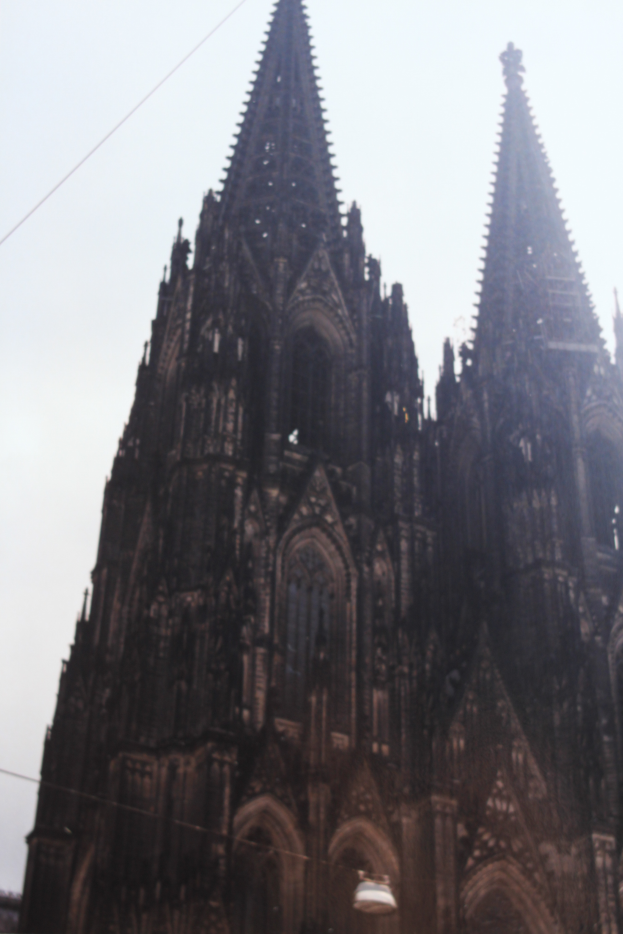 Der Kölner Dom in Köln, Deutschland, mit seinen Türmen, die in den Himmel ragen, und einigen Menschen vor dem Gebäude mit sichtbaren Drähten im Hintergrund.