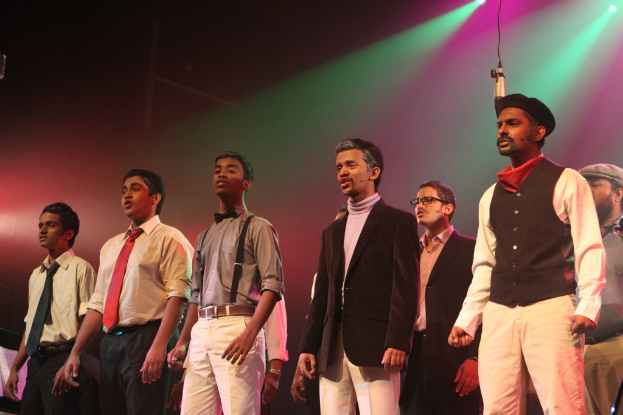 Eine Gruppe von Männern, die auf einer Bühne stehen und ein Drama performen, mit grünen Scheinwerfern im Hintergrund.