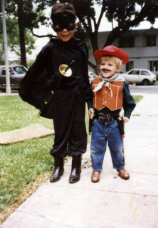Kinder in verschiedenen Kostümen auf einer Straße mit Gebäuden, Bäumen und Fahrzeugen im Hintergrund.