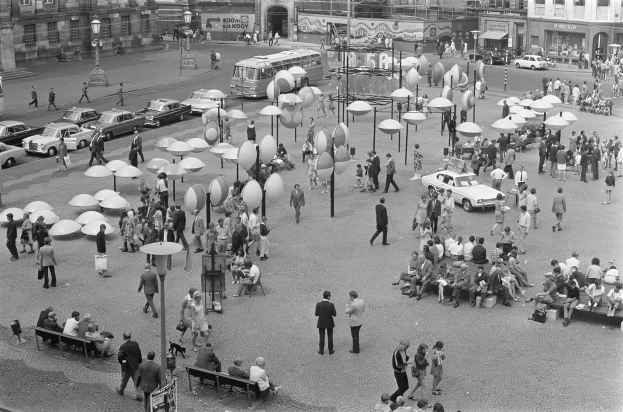Ein Schwarz-Weiß-Foto eines belebten Stadtplatzes mit Menschen, die gehen, auf Bänken sitzen, Laternen und Fahrzeugen sowie Gebäuden mit Fenstern und Namensschildern im Hintergrund.