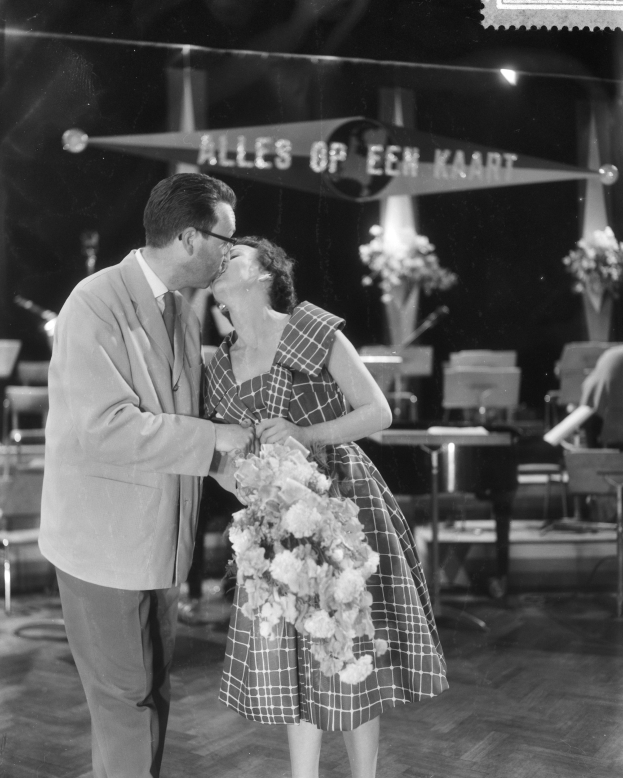 Ein Mann und eine Frau umarmen und küssen sich auf einer Bühne, während die Frau einen Blumenstrauß hält, mit Stühlen und Vasen im Hintergrund.