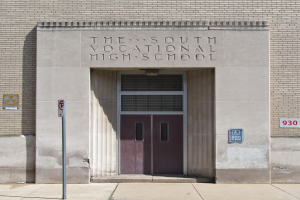 Das Bild zeigt den Eingang der Südberufsschule mit einer Tür, Schildern an der Wand und einem Weg, der hinaufführt.