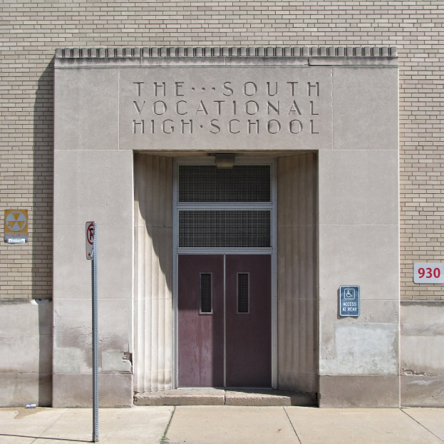 Das Bild zeigt den Eingang der Südberufsschule mit einer Tür, Schildern an der Wand und einem Weg, der hinaufführt.