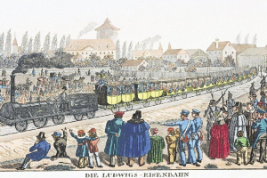 Eine Gruppe von Menschen, die um und in einem Zug auf einem Gleis stehen, mit Gebäuden und Bäumen im Hintergrund und dem Text "Ludwigs-Eisenbahn, der erste Bahnhof in Deutschland" unten.
