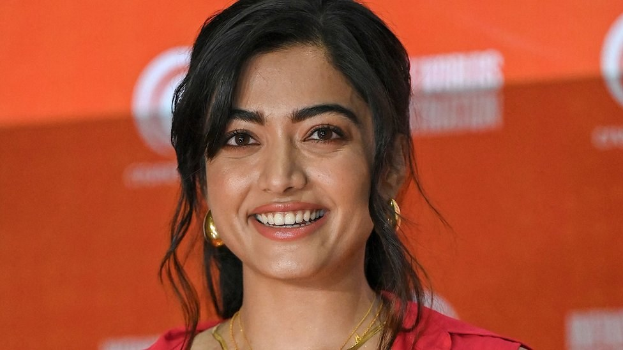 Eine Frau in einem roten Kleid mit einem eleganten Dutt, schwarzen High Heels und natürlichem Make-up lächelt in die Kamera, vor einem hellgelben Banner mit der Aufschrift 'Sonam Kapoors Schönste Looks vom Cannes Film Festival.'